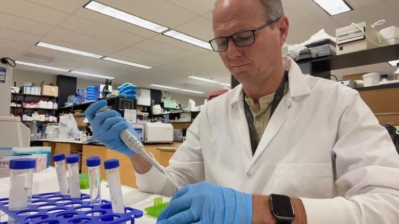 A professor wearing a white lab coat and black glasses perfoms an experiment while sitting down. 