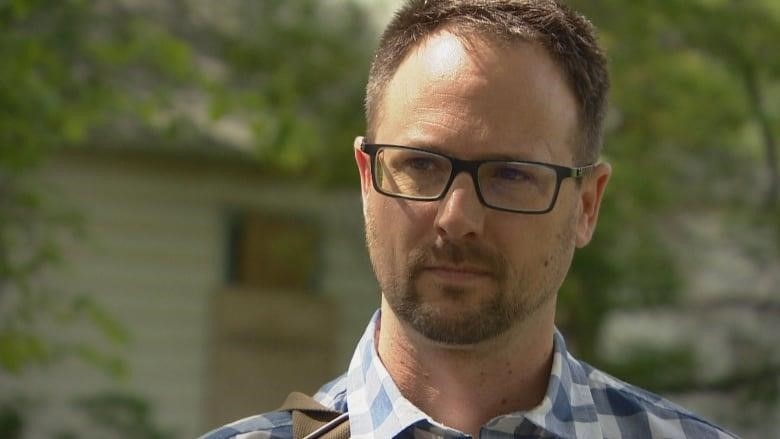A white man with short dark hair, a short beard and glasses is wearing a blue plaid button-up short sleeve shirt. Behind him is a house with a window boarded up.