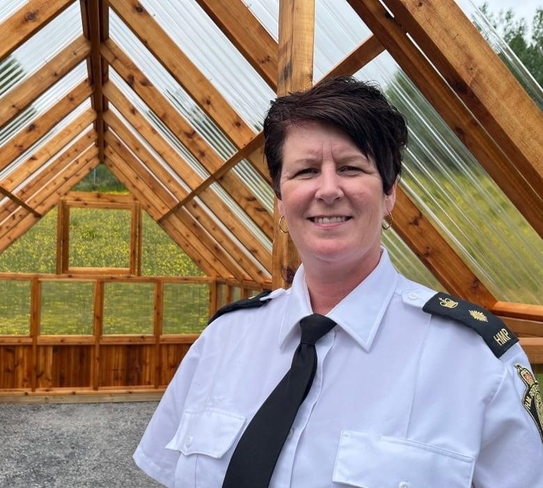 A woman, wearing a uniform, smiles. She stands at the entrance of a greenhouse, which is empty inside.