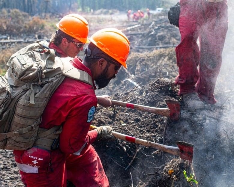 A line of men wearing red firefighting suits walk along a forest path.