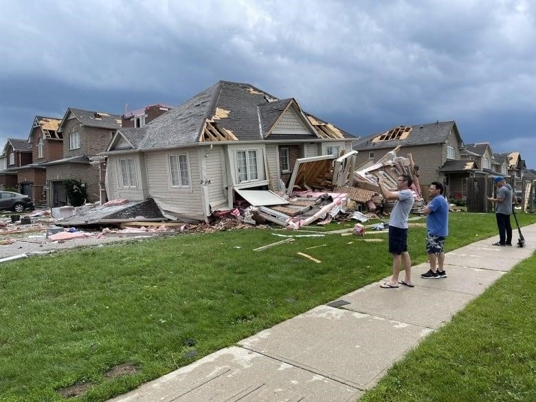 People stand on a sidewalk looking at a damaged home in Barrie. One man takes a photo. 