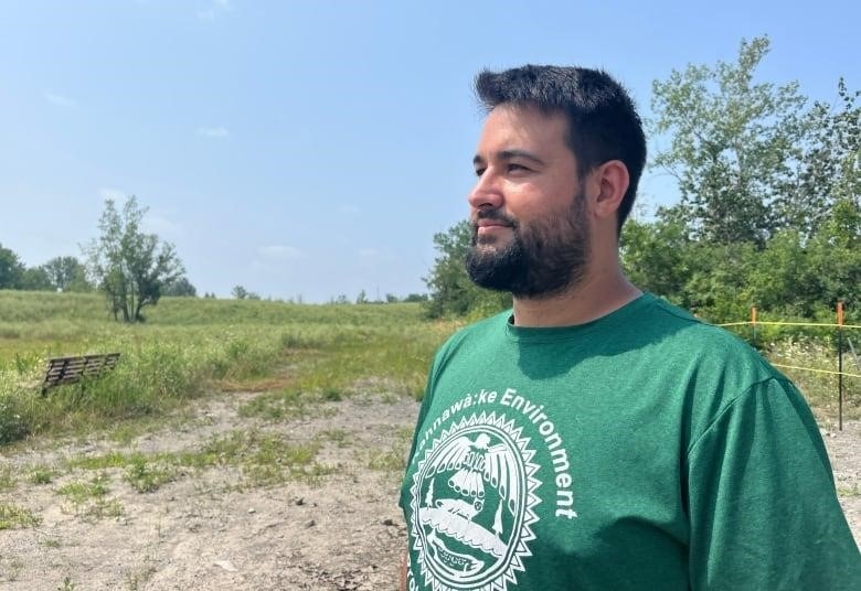 A man wearing a green shirt that says Kahnawà:ke Environment on it looks out onto a marshy bay.