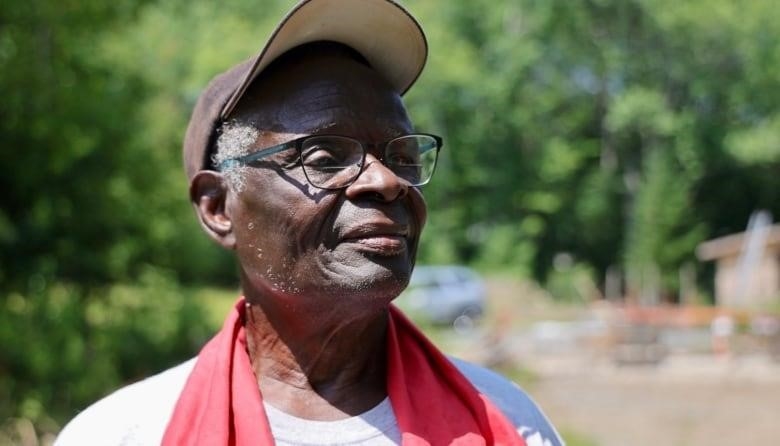 An older Black man with glasses, a ballhat and red cloth around his neck looks to the right side of the frame.