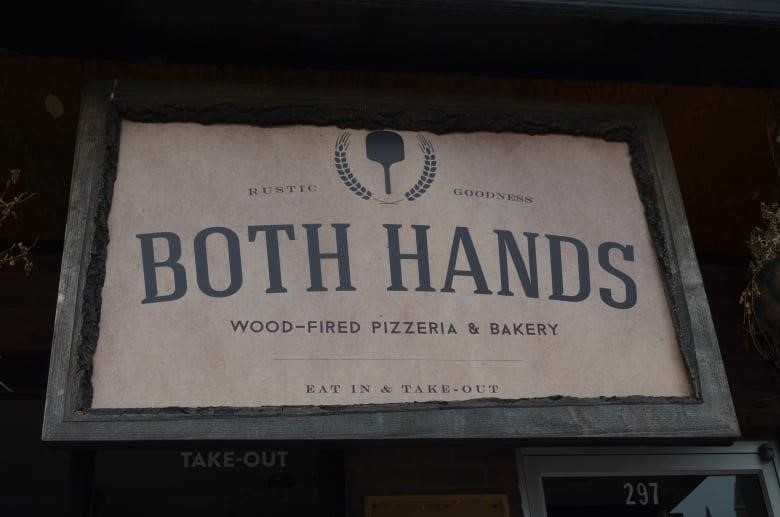 A hanging beige and wooden sign reads "Both Hands wood-fired pizzeria and bakery.:
