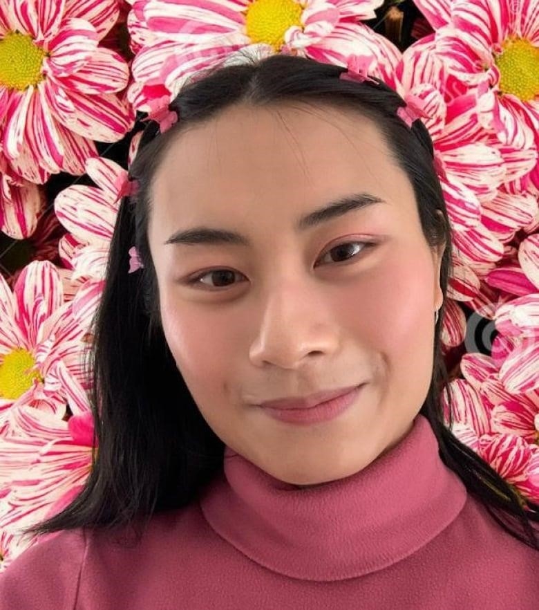 A woman with black hair smiles in a selfie. There are pink flowers in the background. 