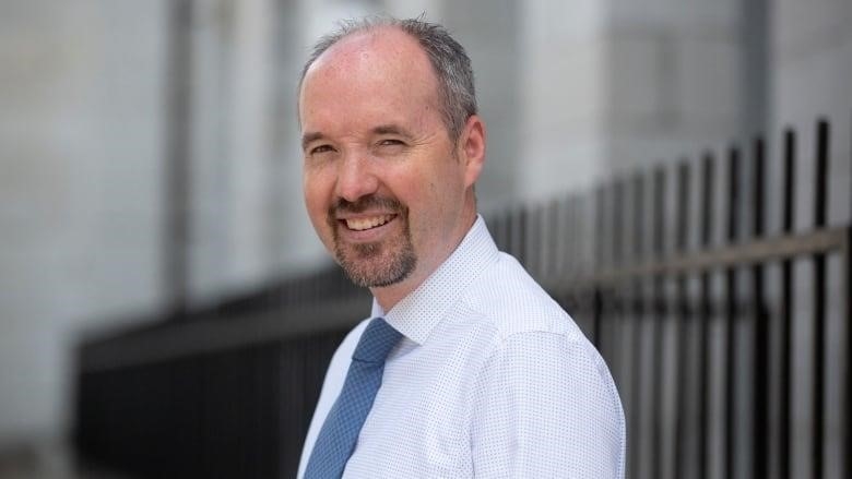 A man with a greying, balding head of hair and a black goatee smiles at the camera. He's wearing a blue shirt and tie and standing next to a black, metal fence.