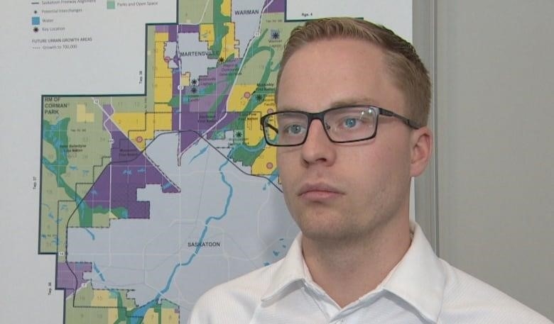 A white man with strawberry-blonde hair is wearing glasses and a white polo shirt. He is standing in front of a map of Saskatoon.