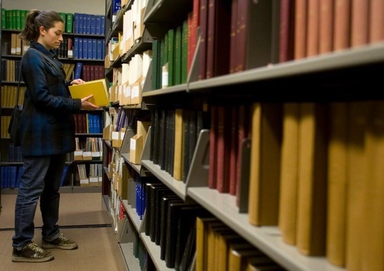 A woman in a blue coat looks at a library book inside a university library. 