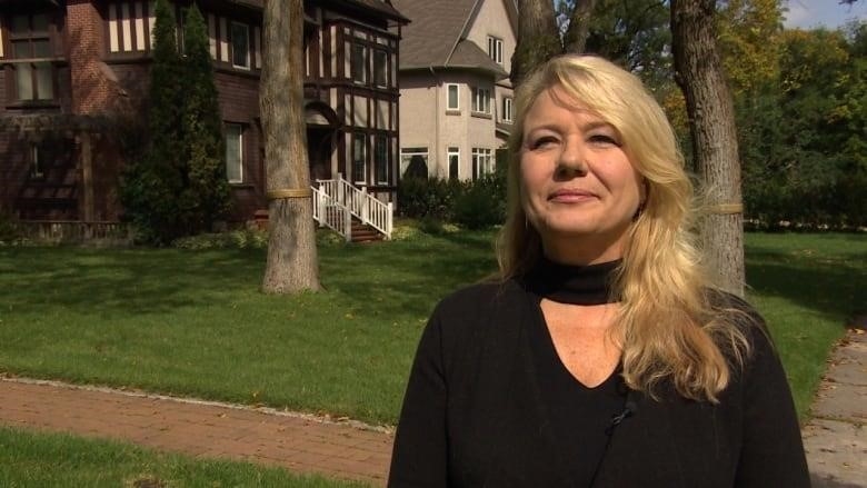 A blonde woman wearing a black shirt stands outside several large houses.