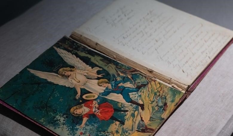 A notebook is shown open on a pillow. The first page shows an angel mother overlooking two children, and cursive writing is shown on the other page.