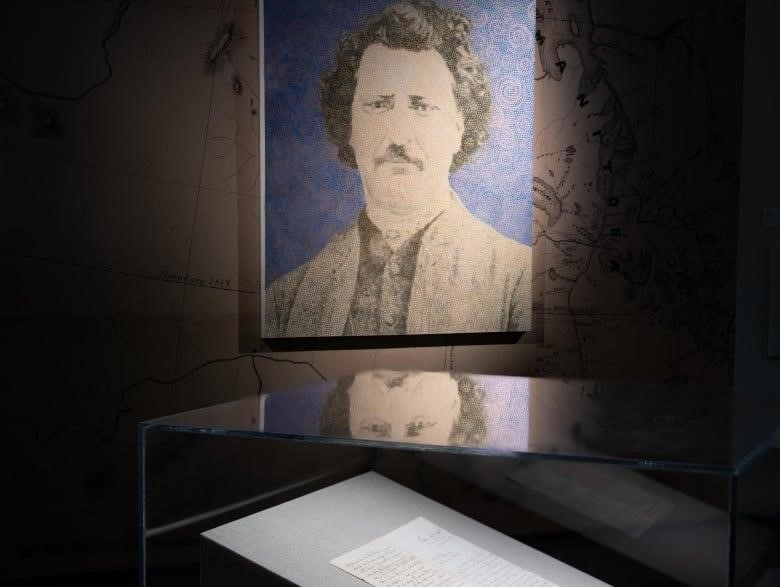 A portrait is shown lit in the middle of a darkened room. It is encircled by letters encased in glass, and quotes painted on the walls, which are also lit.