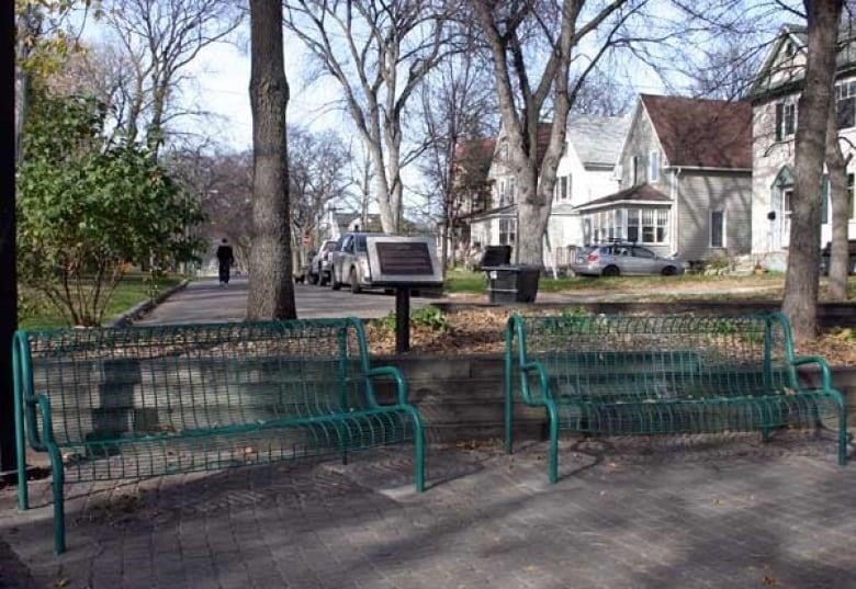 Two green park metal benches are seen with a plaque between them and houses along a street in the background.