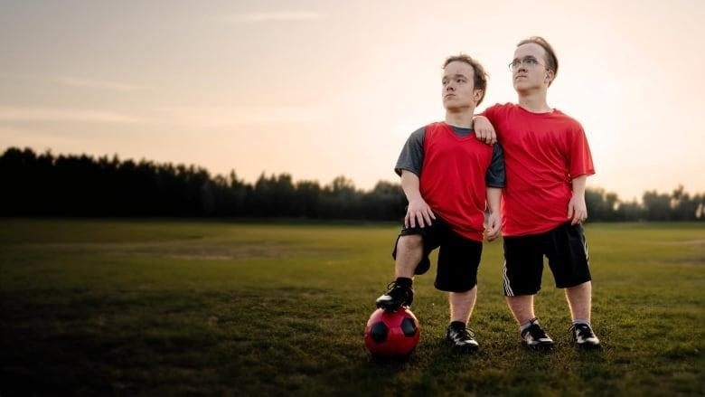 Twins standing together in a field. One of them has their foot on a soccer ball. 