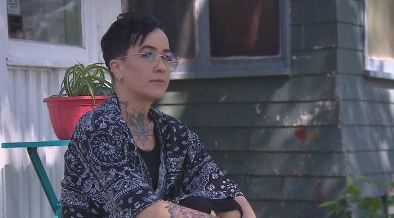 A light-skinned woman with a pixie cut is wearing glasses. She has tattoos on her sternum, neck and right forearm. She is wearing a dark blue shawl with a white pattern on it, while sitting on her front step, in front her wooden house.