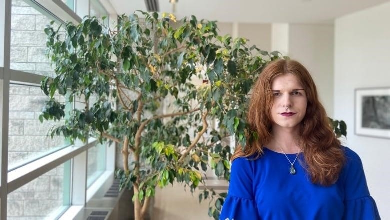 Orange haired trans woman in blue dress stands in front of tree at city hall. 