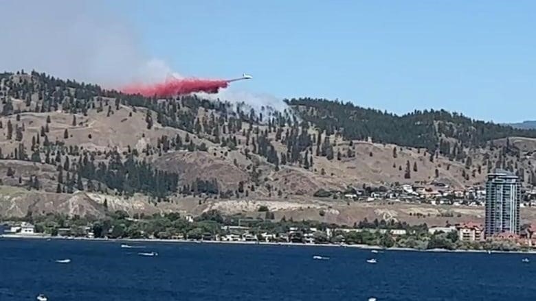 Red plumes of fire retardant drop from an airplane onto smoke from a wildfire on a mountain above houses, an apartment tower and a lake.