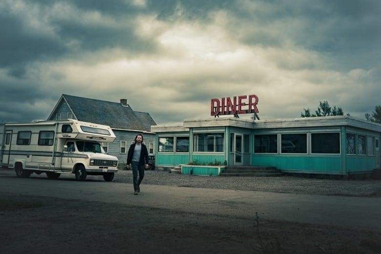 An actor stands in front of a diner on a film set. The sky is cloudy.