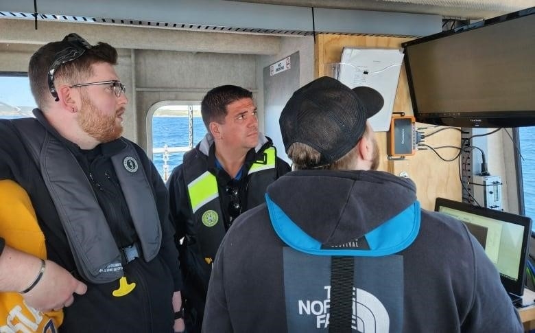 Three men inside a boat stare at a mounted computer monitor.