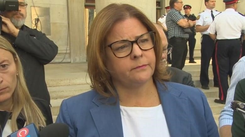 A woman wearing glasses and a blue blazer speaks to reporters holding microphones.