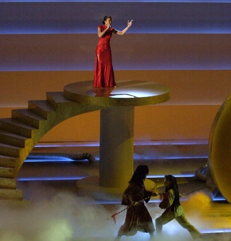 A woman on stage in a red dress holds a microphone at the top of a flight of stairs ending in the small landing she is standing on.