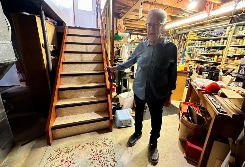 A man stands on a basement floor beside the staircase, looking up the staircase at the camera as he holds on to the handrail.