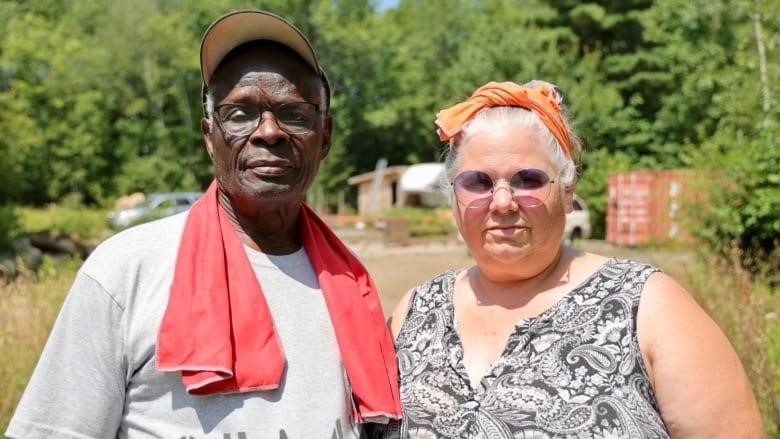 An older couple, Black man and white woman, stand together looking straight at the camera. They are outside in a treed area.