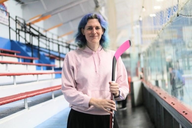 A woman with dyed hair is wearing a hoodie and holds a hockey stick in a hockey arena.