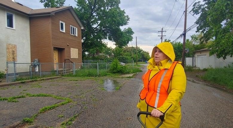 A light-skinned woman is wearing an orange safety vest over a yellow raincoat. Her hood is up. She is holding the handle of a wagon with her left hand. She is standing on asphalt, in front of two housing units with placarded windows and doors. A large tree stands behind the residences.