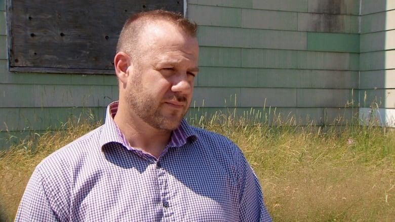 A man stands in front of a boarded up home. 