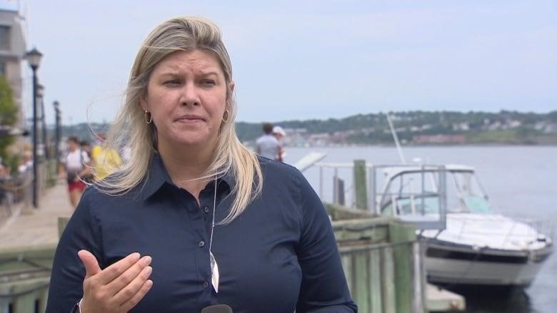A woman in a blue jacket speaks in front of boats and water.