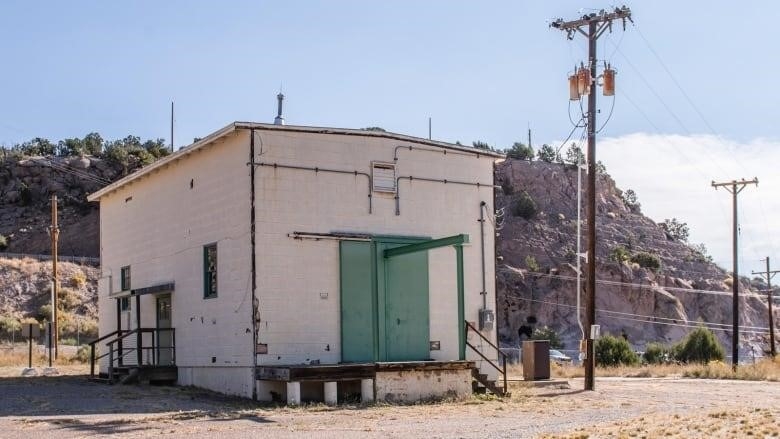 A industrial-looking white building with green doors stands in the foreground. A range of hills runs behind it.