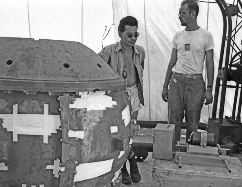 Two men, one in dark sunglasses, stand near a large metal sphere.