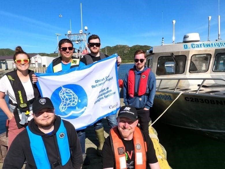 5 men and 1 woman pose on a wharf aside a silver boat. A man in the center of the huddle holds up a flag that reads' Ghost Gear Retrieval Project.'