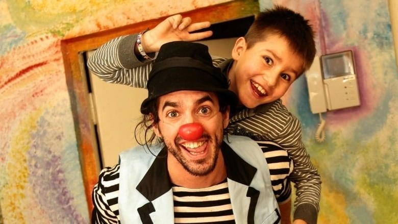 A man dressed in blue and a red clown nose smiles at the camera with a kid on his back, smiling. 