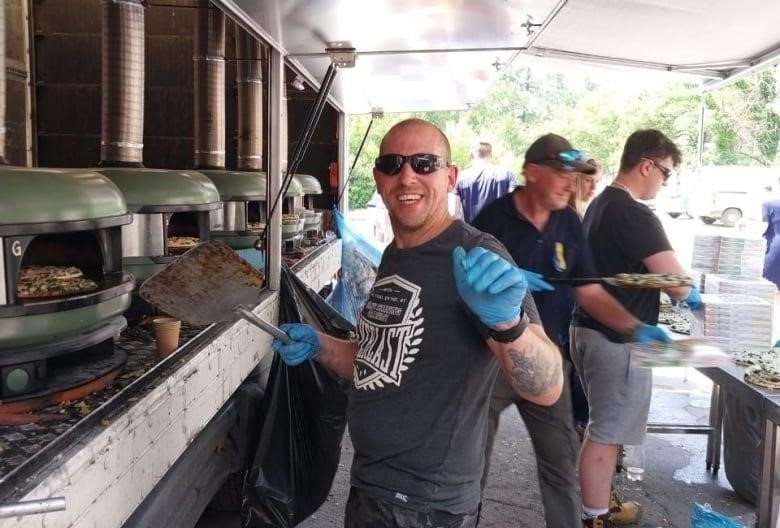 A man smiles at the camera while watching over several pizza ovens that are in the side of a truck. 