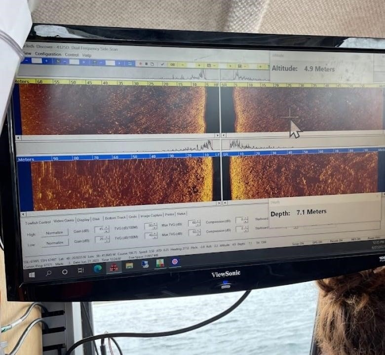 A computer monitor shows four crude maps of the ocean floor. The monitor is suspended above a window showcasing the ocean outside the boat.