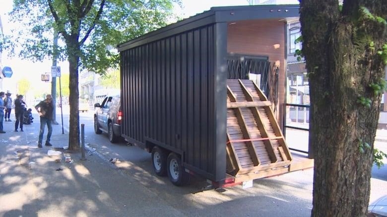 A small black trailer parked on the street with a wooden ramp in the back.