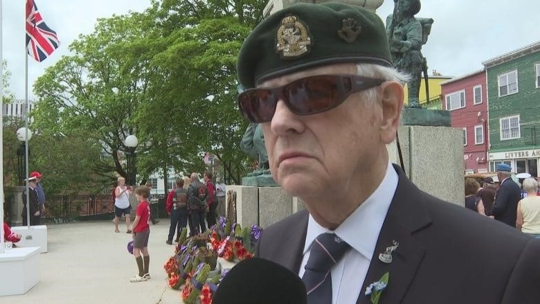 A man in sunglasses and a military beret looks grim in front of the National War Memorial in St. John's.