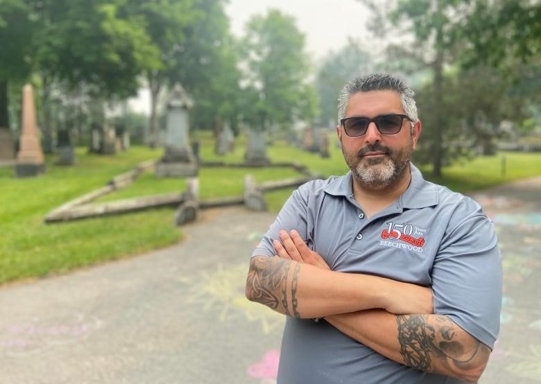 Man with sunglasses stands in cemetery with arms crossed