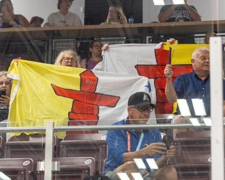 Several people sit in a stadium behind plexiglass, holding up bright flags.