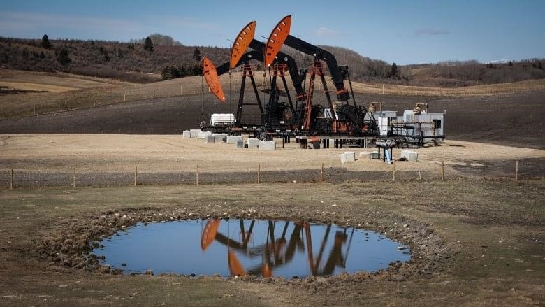 An oil pumpjack is pictured in front of a body of water, with the pumpjack's reflection displayed in the water. 