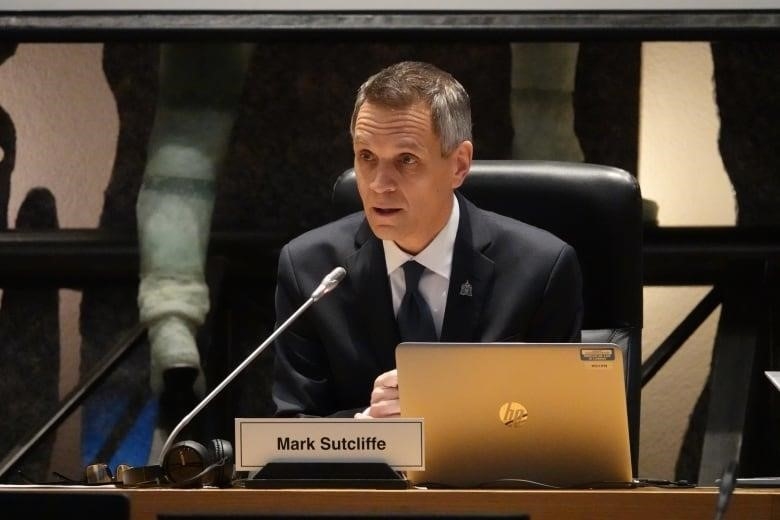 A man in a suit sits in front of a microphone and a laptop and delivers a speech.