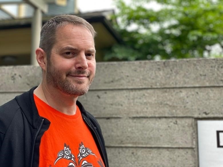 A person stands in front of a wall, with trees behind.