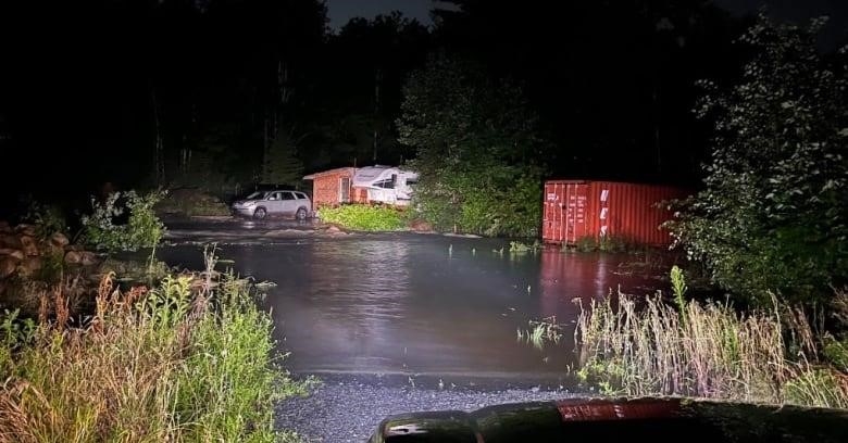 A nighttime photo shows a large grey pool of water forming in front of a gravel driveway, blocking in an RV partially obscured by trees