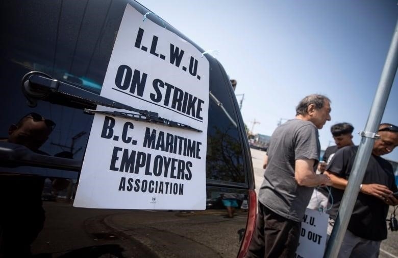 A sign reading "I.L.W.U workers on strike" is affixed to the back of a black SUV, and a group of three men holding similar signs and looking down at their phones stand next to the vehicle.