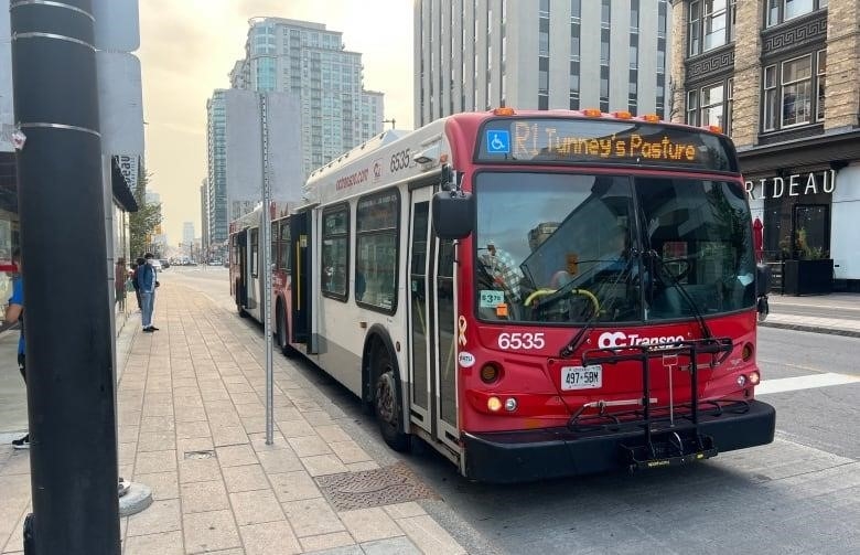 A bus that says R1 Tunney's Pasture stops on a street outside a downtown mall in summer.