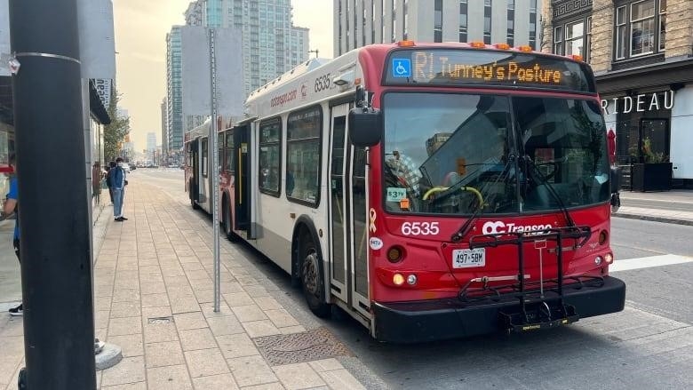 A bus that says R1 Tunney's Pasture stops on a street outside a downtown mall in summer.