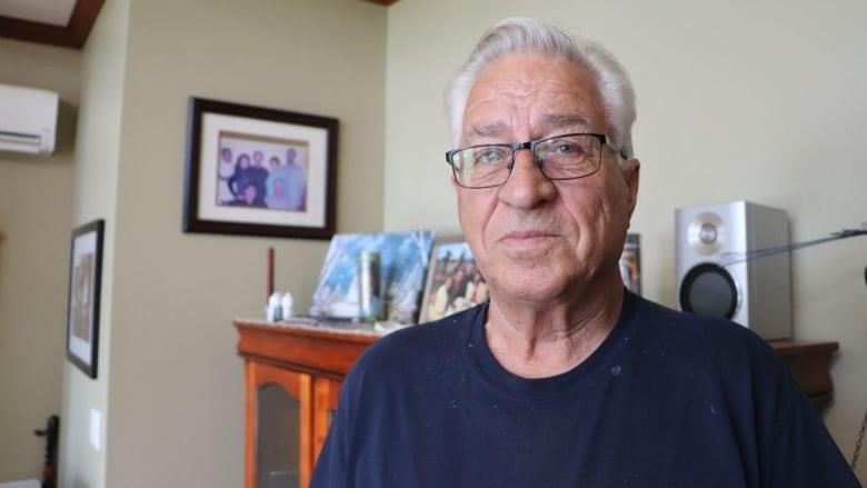 A man looks at the camera. Behind him are framed family photos. 