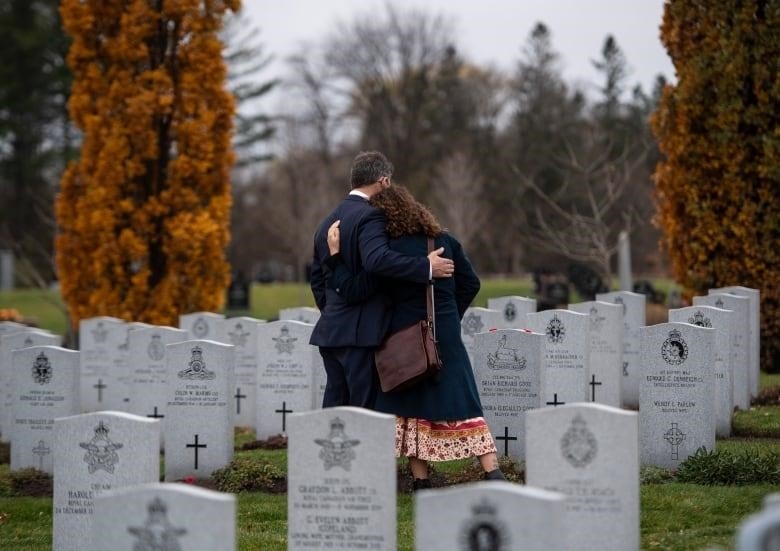 Two people are seen from behind, comforting each other, while standing amidst dozens of graves.