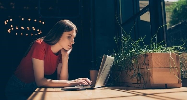 A woman sits at a table, looking at a laptop open in front of her.